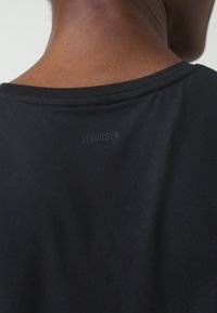 Gros plan sur le haut du dos d'une personne portant un t-shirt noir avec le mot « STRONGER » imprimé en petites lettres foncées sous l'encolure.