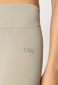 Világos bézs leggings, bordázott derékpánttal, sima textúrával és diszkrét zöld logóval. Az anyag ellentétes mintázatú résszel rendelkezik.