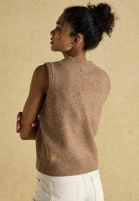 Brauner ärmelloser Strickpullover mit strukturiertem Finish und geripptem Halsausschnitt; getragen über hellen Hosen, die ein dezentes Rückendetail zeigen.