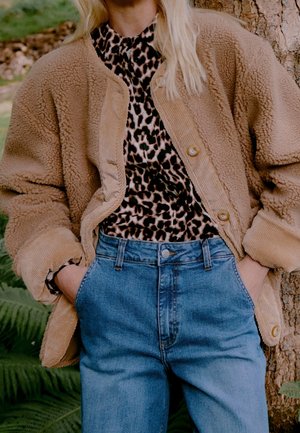Osoba ubrana w beżową kurtkę sherpa na leopardzianej koszuli, z rękami w kieszeniach niebieskich, wysokich dżinsów, stojąca przy pniu drzewa.