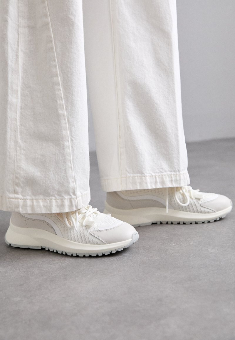 White sneakers feature a textured upper with mesh and smooth leather accents, a rounded toe, and a chunky sole with black tread.