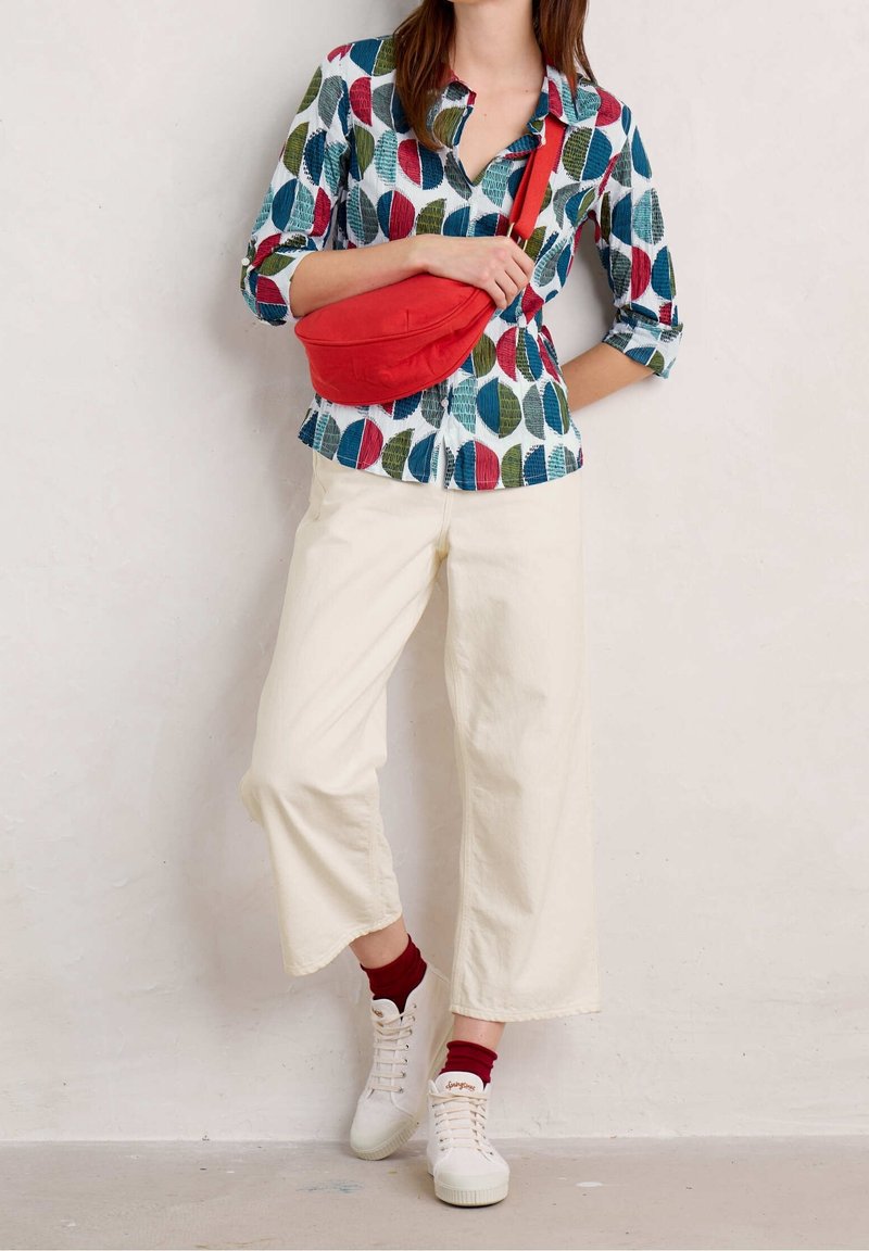 Colorful leaf-patterned shirt in blue, green, and red, white background; paired with cream wide-leg pants and red-accented sneakers. Red bag.