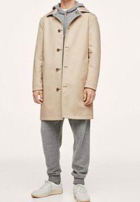 Beżowy płaszcz przeciwdeszczowy z kapturem, zapięcie na guziki i boczne kieszenie, nałożony na szary sweter i dopasowane dresowe spodnie, z białymi sneakersami.