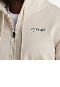 Felpa beige con zip realizzata in misto cotone. Presenta una zip frontale, cappuccio con cordino e il logo blu ricamato "G-Star Raw" sul petto.
