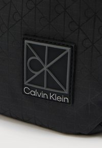 Μαύρη υφασμάτινη τσάντα με υφή σχεδίασης. Διαθέτει ορθογώνιο ελαστικό έμβλημα με την επιγραφή "Calvin Klein" και γεωμετρικά σχέδια σε γκρι.