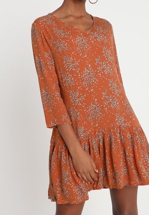 Vrouw die een losse, roestoranje jurk draagt met witte bloemenpatronen, driekwartmouwen en een gerimpelde zoom.