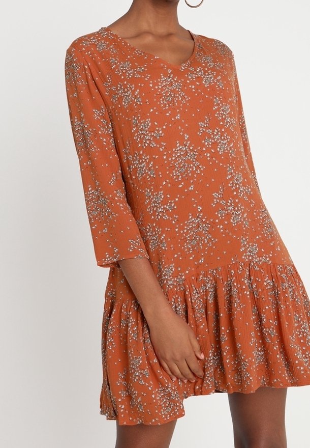 Femme portant une robe ample couleur orange rouille avec des motifs floraux blancs, manches trois-quarts et un ourlet à volants.