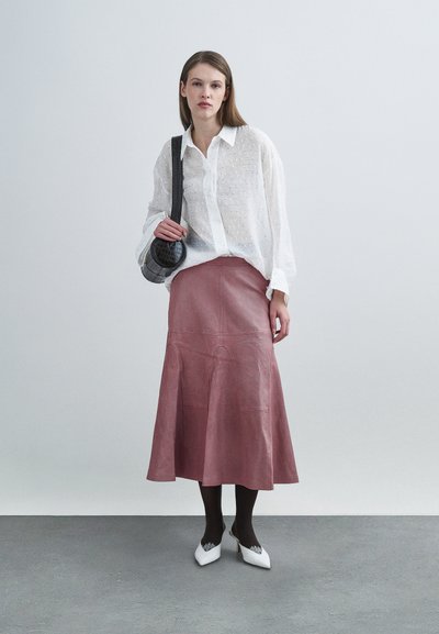 Blouse blanche texturée à manches longues, associée à une jupe évasée en cuir rose. Le mannequin porte des chaussures à talons blanches et tient un sac noir.