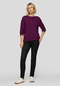 Lila gestreifter Strickpullover mit langen Ärmeln, kombiniert mit schwarzen Jeans und weißen Sneakern. Das Model steht vor einem neutralen grauen Hintergrund.