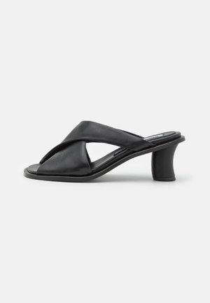 Sort åben tå mule sandal med brede polstrede krydsede stropper og en buet, mellemhøj blokhæl på en hvid baggrund.