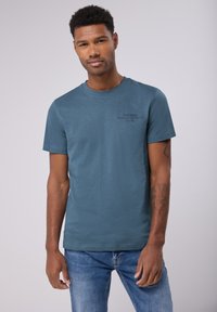 Blå kortärmad t-shirt i mjukt material, med rundad halsringning och en tryckt grafik i svart på övre vänstra sidan.