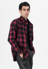Camisa de cuadros rojos y negros en una tela suave con cuello abotonado y mangas largas, combinada con pantalones negros texturizados.