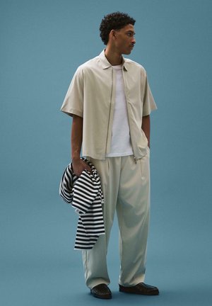 Férfi bézs rövid ujjú cipzáras dzsekiben és nadrágban, fehér inggel alatta, kék háttér előtt fekete-fehér csíkos összecsukott pulóvert tartva.
