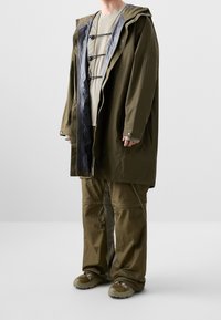 Personne portant un imperméable à capuche vert olive avec un pantalon assorti et des chaussures de randonnée, debout devant un fond clair uni.