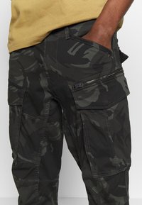 Donkere camouflage cargobroek met ritszakdetail, gedragen door een persoon met één hand in de voorzak en een mosterdgele shirt.