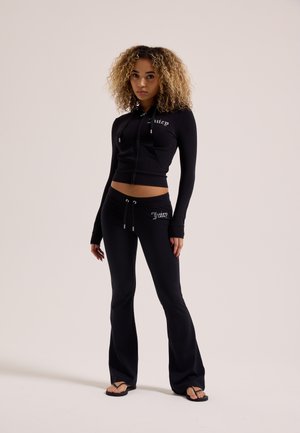Donna con capelli ricci, indossa una felpa nera corta con zip e pantaloni neri svasati abbinati con il logo "Juicy", in piedi con infradito nere.