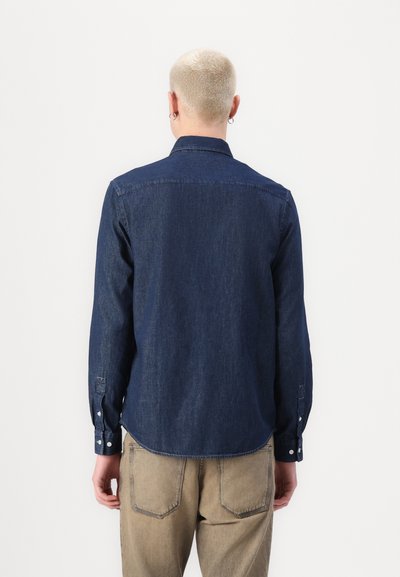 Chemise en denim bleu foncé à manches longues, avec des poignets boutonnés et un col classique, présentant une texture lisse et un ourlet légèrement arrondi.