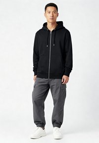 Giovane uomo in piedi che indossa una felpa nera con zip, pantaloni cargo grigi e sneakers bianche su uno sfondo chiaro e uniforme.