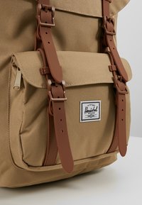 Khaki plecak z kanwy z brązowymi skórzanymi paskami, przednią kieszenią i łatą z logo. Posiada zapięcie na klapkę oraz teksturowany materiał.