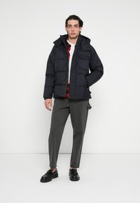 Granatowa pikowana kurtka puchowa z kapturem, wyposażona w pikowane wypełnienie, przednie kieszenie i zapięcie na guziki. W połączeniu z szarymi spodniami i czarnymi butami.