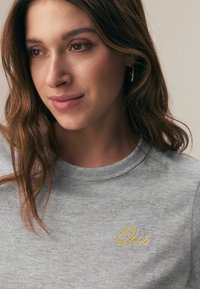 Siva majica s kratkimi rokavi, z okroglim izrezom in rumeno vezenim napisom "Oui" na prsih. Mehka tekstura tkanine.