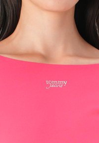 Rozā, pieguļošs tops ar plecus atstarojošu dizainu un metālisku sudraba "tommy jeans" logotipu kreisajā pusē. Gluda tekstūra.