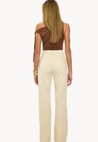 Top marrón asimétrico con detalle de lazo, combinado con pantalones de tiro alto y pernera ancha en color beige. Pulsera grande de oro en la muñeca izquierda.