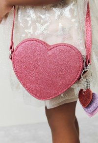 Růžová crossbody taška ve tvaru srdce vyrobená z třpytivého materiálu, s popruhem a přívěskem, které obsahují menší srdíčka v červené a fialové barvě.