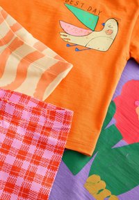 Orange kortærmet skjorte med en fuglegrafik og teksten "BEDSTE DAG". To mønstrede stofstykker: et stribet i creme og orange, og et pink og rødt ternet.