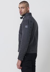 Sötétszürke farmerdzseki magas gallérral, steppelt vállrésszel, oldalsó cipzáras zsebekkel és textúrált felülettel. A ujjon logó folt található.