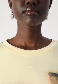 Light yellow t-shirt with a soft texture, featuring a round neckline and subtle logo detail. Silver-colored drop earrings complement the look.