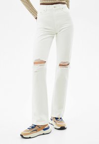White high-waisted jeans with ripped knees; made of denim with a smooth texture. Paired with multicolored sneakers featuring mesh and leather accents.