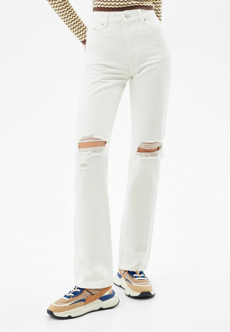White high-waisted jeans with ripped knees; made of denim with a smooth texture. Paired with multicolored sneakers featuring mesh and leather accents.