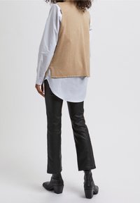 Beige gebreide vest over een wit shirt met een gebogen zoom, gecombineerd met zwarte leren broeken en enkellaarsjes, bekeken van achteren.