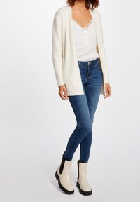 Ljusbeige fluffig cardigan över en vit topp med spetskant, i kombination med blå skinny jeans och krämfärgade ankelstövlar med svarta detaljer.
