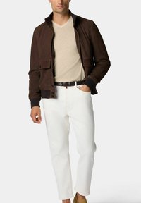 Braune Canvasjacke mit zwei Fronttaschen, beigem V-Ausschnitt-Shirt, weißen Jeans und hellbraunen Schuhen. Die Jacke hat einen Druckknopfverschluss.