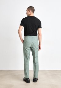 Chemise noire à manches courtes associée à un pantalon ajusté vert clair au tissu lisse et doté de deux poches arrière, portée avec des chaussures noires.