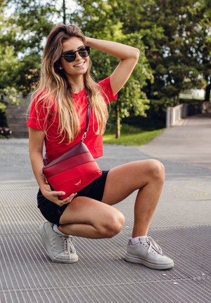 Rot- und dunkelroter Umhängetasche mit horizontalen Streifen, aus glattem Leder. Das Modell trägt ein rotes Hemd und schwarze Shorts, auf einer gemusterten Oberfläche.