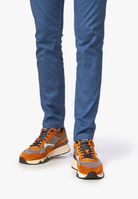 Sneaker multicolori con suede arancione, rete grigia e una spessa intersuola color crema. Lacci neri con occhielli in metallo. Abbinati a pantaloni blu.