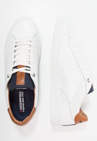 White sneakers with leather upper, textured sides, and light brown heel accent. Features round toe and white laces over a navy blue tongue.