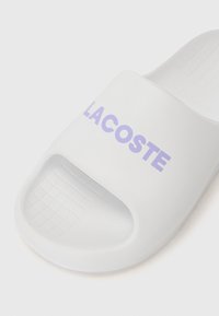 Sandale blanche à glissière en matériau lisse avec un logo "LACOSTE" violet. Design ouvert à l'avant avec une semelle texturée pour une bonne adhérence.