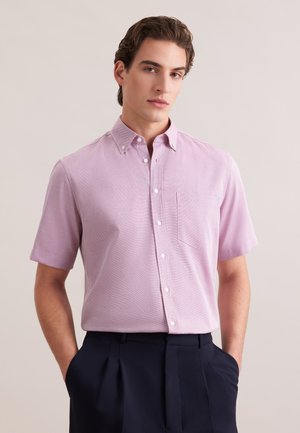 Młody mężczyzna ubrany w jasnofioletową, krótkorękawową koszulę zapinaną na guziki z kieszenią, wpuszczoną w ciemne spodnie z plisami, z rękami w kieszeniach.