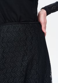 Zwarte kanten rok met bloemenpatroon, voorzien van een effen zwarte top en een elastische tailleband. De rok heeft een gestructureerde, doorschijnende kanten afwerking.