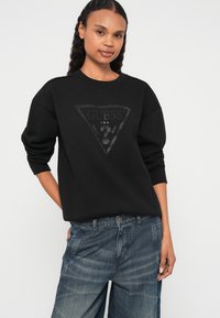 Černá mikina s texturovaným logem ve tvaru trojúhelníku na přední straně, volnější střih a žebrované manžety. Spárovaná s vybledlými modrými džínami.