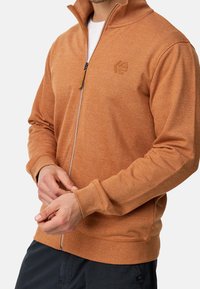 Sweatshirt orange à zip fabriqué en tissu texturé. Il possède un col montant, des poignets côtelés et un petit logo sur la poitrine gauche.