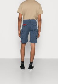 Blauwe cargoshorts met zijzakken, verstelbare manchetten en patchdetails, gecombineerd met een beige T-shirt en zwarte hoge sneakers.