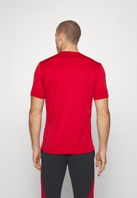 Homme aux cheveux courts portant une chemise rouge à manches courtes et un pantalon noir et rouge, de dos contre un fond clair uni.
