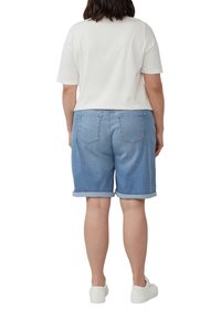 Person trägt ein weißes kurzärmeliges Hemd, helle, hochgerollte Jeansshorts und weiße Turnschuhe, steht mit dem Rücken zur Kamera.