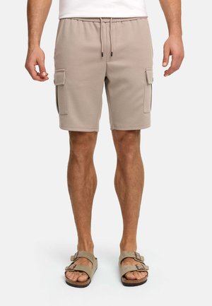 Mann trägt beige Cargoshorts mit Frontklappentaschen, Taillenbund mit Kordelzug und beigefarbenen Schnallensandalen vor weißem Hintergrund.