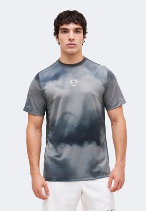 Młody mężczyzna ubrany w szarą i czarną sportową koszulkę z krótkim rękawem z wzorem chmur i małym białym logo Swoosh na piersi.